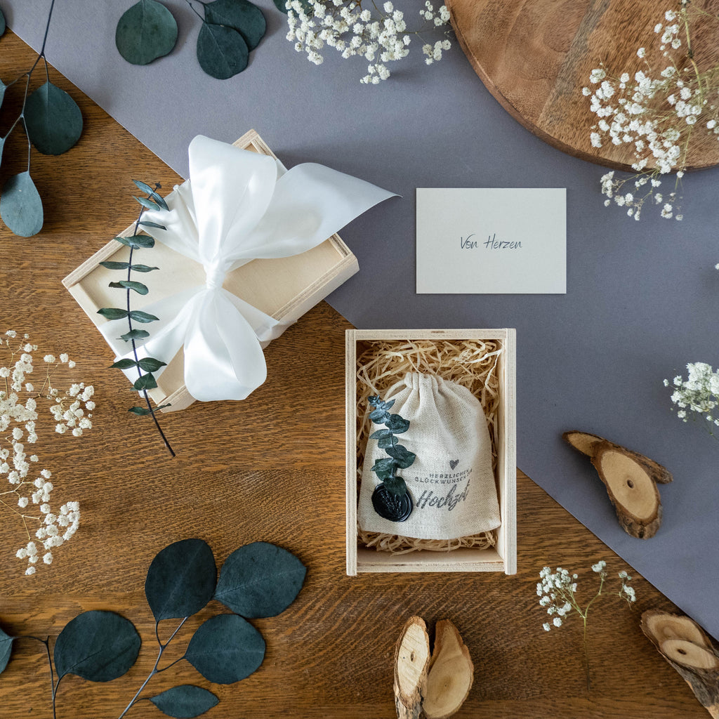 Holzbox mit Schiebedeckel und weißer Satinschleife, gefüllt mit Jutebeutel "Herzlichen Glückwunsch zur Hochzeit" und Karte "Von Herzen".